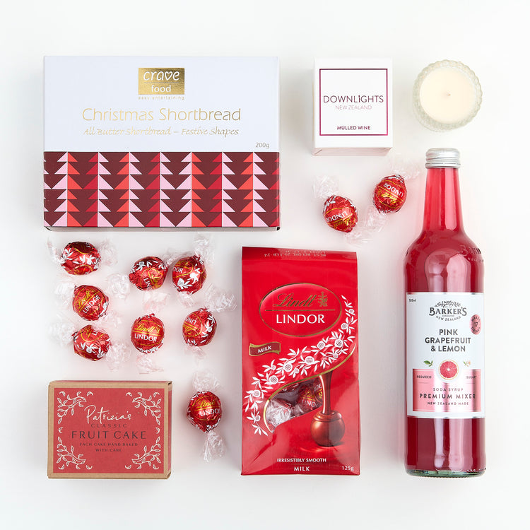 A collection of gift items including a red candle, a box of Lindt chocolate, a bottle of Barkers mixer, wrapped candies, a box of Christmas shortbread, and a box of Patrizia fruit cake.