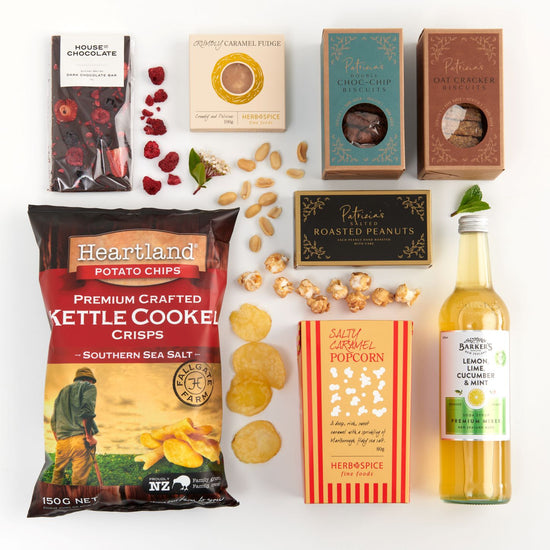 A collection of food items including chocolate bars, potato chips, biscuits, roasted peanuts, crackers, popcorn, and a bottle of spritzer arranged on a white surface.