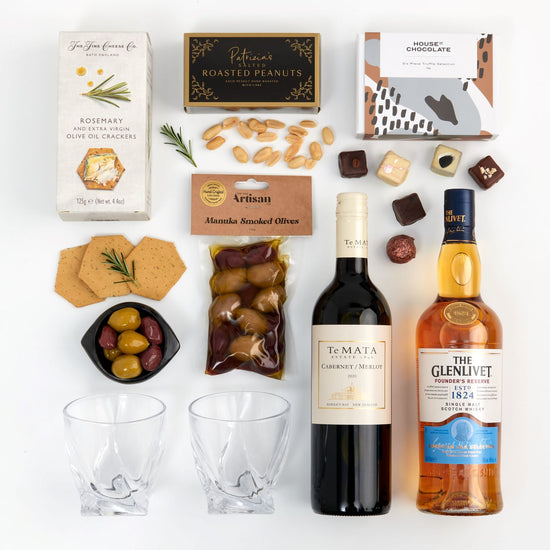 A gift box presentation containing alcohol bottles, whisky tumblers, crackers, smoked olives, and chocolate truffles.